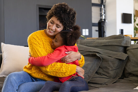 Hugging Diverse mother and daughter cuddling on grey sofa in living room, with modern fireplace