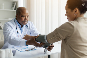 Obraz premium Mature Black Doctor Man Checking Blood Pressure To Female Patient With Upper Arm Monitor, Professional Physician Male In Uniform Measuring Arterial Tension To Young Woman During Appointment In Clinic