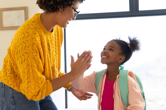 High-fiving Diverse mother and daughter indoors near large window, with mint green backpack