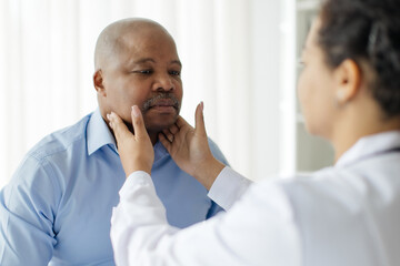Medicine and healthcare concept. Black female doctor checking lymph nodes of male patient at hospital, carefully examining the neck and throat area of mature man in bright medical clinic setting
