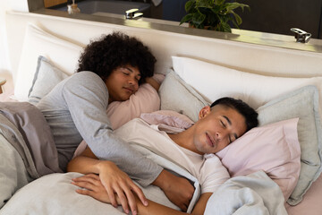 Cuddling Diverse male partners resting on bed in bedroom, with pink pillows and potted plant