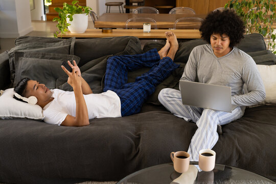 Lounging Diverse male friends scrolling smartphone and typing on laptop in living room, with mugs
