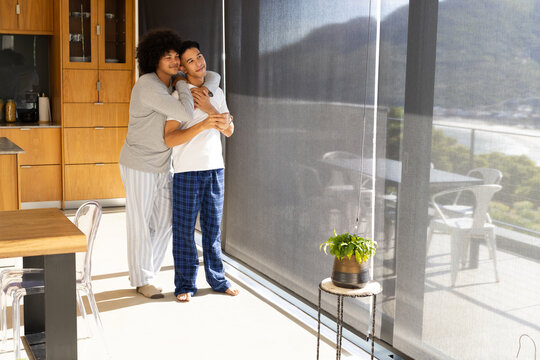 Hugging diverse male duo standing near sliding glass doors at modern apartment, with balcony view