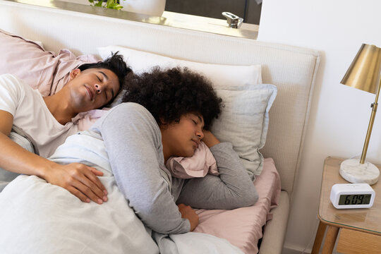 Sleeping diverse male partners under gray duvet in modern bedroom, with digital alarm clock