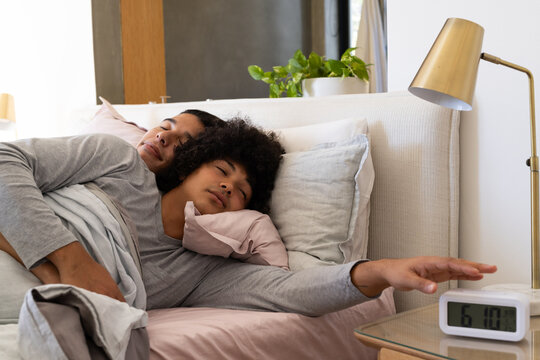 Diverse male partners lying and reaching in cozy bedroom, with digital alarm clock on bedside table