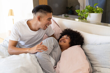 Smiling Diverse male partners leaning over each other in bright cozy bedroom, with pink duvet