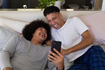 Laughing diverse male partners leaning on headboard in bedroom-bathroom space, with smartphone