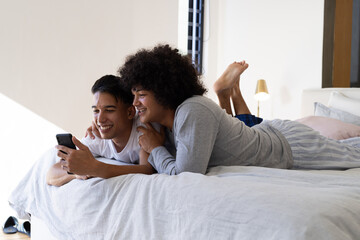 Lying diverse male partners cuddling on bed at home holding smartphone with pastel pillows and lamp