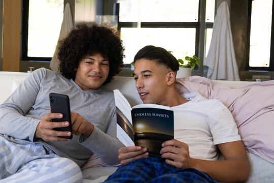 Lounging Diverse male friends reading Endless Sunsets and checking smartphone on bed with blanket