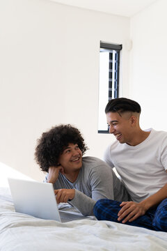Laughing diverse male partners relaxing on white bed in bright bedroom, with silver laptop