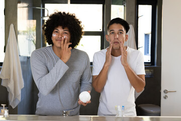 Applying facial cream, diverse male friends standing at bathroom sink, with open jar and mirror