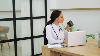 Black female nurse sits at desk in doctor's office working on computer in hospital drinks hot coffee. Mid adult nurse seated at desk medical office working on computer drinking hot cup of coffee. - Powered by Adobe