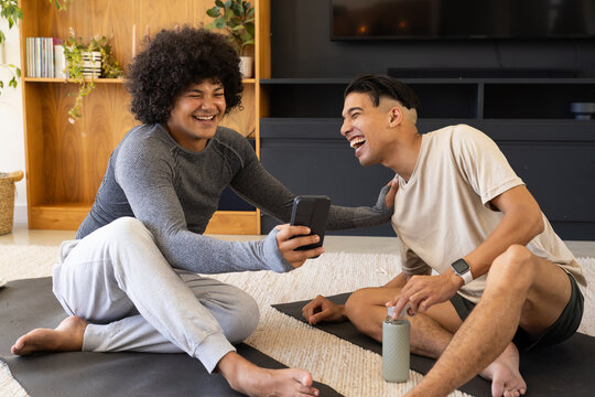 Smiling diverse male friends holding smartphone and water bottle on yoga mats in living room - Powered by Adobe