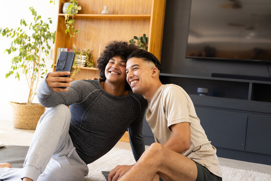 Smiling diverse male friends taking selfie on yoga mat in living room, with smartphone and plants