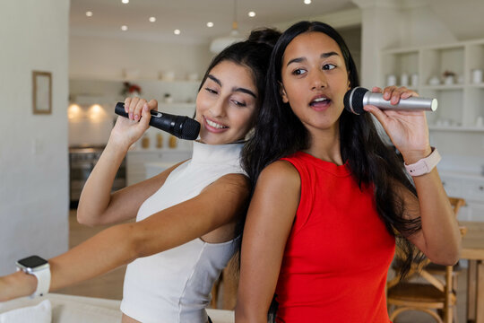 Diverse female friends singing karaoke in modern kitchen, with microphones and smartphone