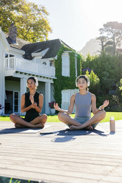 Meditating Diverse female friends sitting cross-legged on backyard deck with mats and headphones