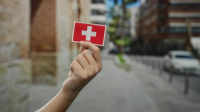 Man holding embroidered swiss flag patch on a city street, highlighting cultural symbolism, identity, and travel appeal in a vibrant outdoor urban setting.