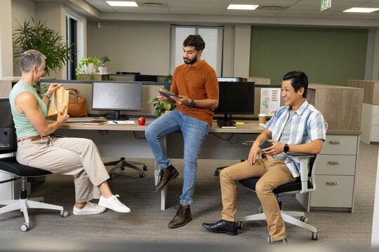 Collaborating Diverse coworkers unpacking lunch bag and tapping tablet with coffee in office
