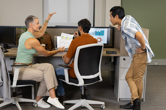Collaborating Diverse coworkers examining data at desk in open-plan office, with monitors, coffee