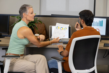 Diverse coworkers reviewing printed report at modern open-plan office desk, with computer monitors