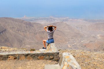Woman sitting on a stone wall at the top of Jebel Jais, enjoying the vast mountain view and coastline below. Scenic, serene, breathtaking moment in Ras Al Khaimah mountains, UAE