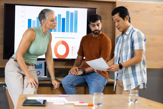Reviewing Diverse coworkers examining printed reports at table in meeting room, with monitor charts