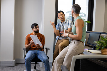 Diverse coworkers discussing printed document at desk in modern office, with pen and monitors