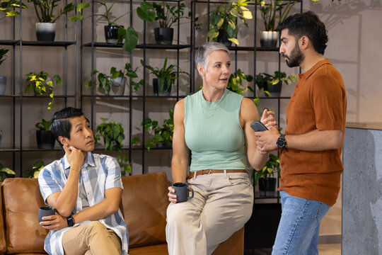 Talking over coffee mugs, diverse coworkers sitting on sofa in office lounge, with smartphone
