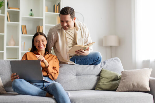 Young couple sitting on couch, spending quality time, woman using laptop, man reading book, both looking at computer screen, copy space