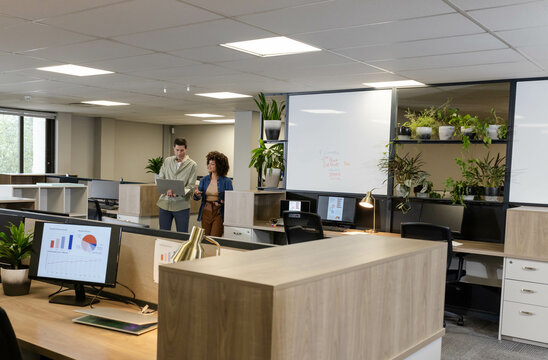 Discussing data, diverse coworkers standing in open-plan office, with laptop and monitor graphs