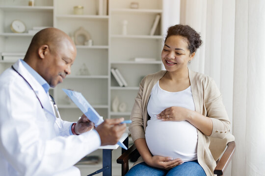 Male Doctor Showing Clipboard With Medical Test Results To Black Pregnant Female Patient During Meeting In Clinic, Gynecologist Man Demonstrating Complete Blood Count Range To Young Smiling Woman