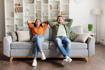 Happy relaxed couple leaning on couch and holding hands behind head, enjoying comfort of their...