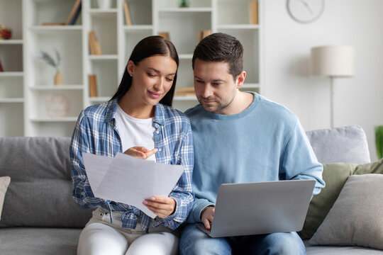 European spouses reading letter from bank, sitting on sofa with laptop, checking correspondence together, copy space. Mortgage, loan concept