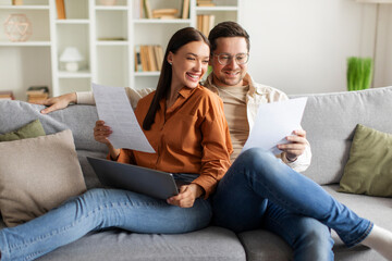 Obraz premium Happy spouses with documents and laptop computer, engaged in discussion and financial planning, sitting on sofa in cozy living room