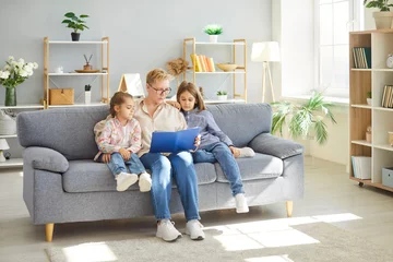 Fotobehang Grandmother and two children sitting on couch, reading interesting book in living room. Happy family spending leisure time together, enjoying fairytale story. Storytelling and child development. © Studio Romantic