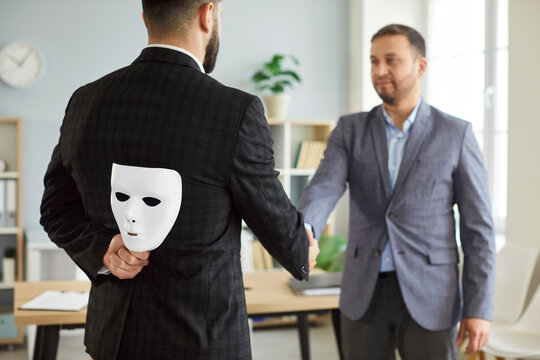 Businessman wearing suit giving dishonest handshake hiding white face mask behind his back. The concept of hypocrite agreement, treachery risk and betray business partnership concept