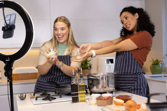 Smiling diverse female friends holding glass mixing bowls at kitchen island, with ring light setup