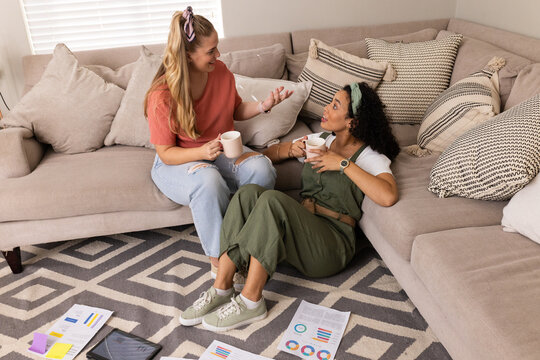 Relaxing Diverse female friends sitting on sofa in living room, with coffee mugs and tablet