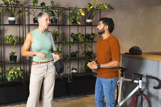 Talking senior woman and Indian man standing in co-working lounge, with bicycle helmet and plants