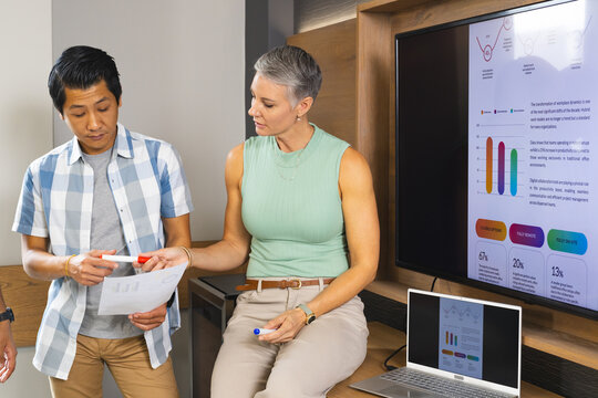 Collaborating Diverse coworkers reviewing printed charts in office corner, with laptop and markers