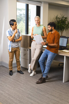 Diverse coworkers reviewing printed diagrams at office desk, with monitor and plant