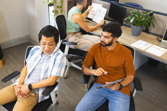 Collaborating Asian male coworkers analyzing data on tablet and printed chart in open-plan office