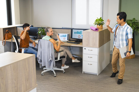 Waving Diverse coworkers sitting at open-plan office desks, holding laptop by dual monitors
