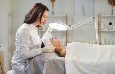 Professional female dermatologist cosmetologist, beautician doing female face exam study under lamp, skin beauty treatment, improving appearance, relaxing girl facial cleansing in beauty care clinic