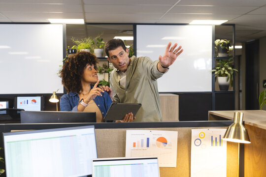 Gesturing diverse coworkers standing at cubicle in open-plan office, with tablet, whiteboard panels
