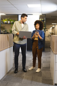 Talking Diverse coworkers standing in office corridor by plants, cubicles, with laptop smartphone