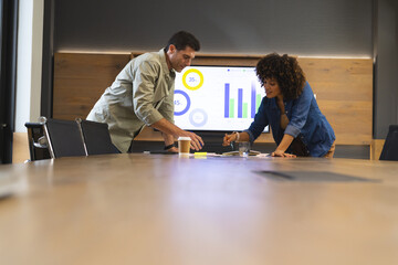 Diverse coworkers leaning over documents at office meeting table, with analytics charts on screen