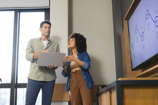 Discussing Diverse coworkers standing in modern office meeting space, with laptop and TV graph