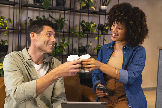 Clinking Diverse coworkers raising coffee cups in lounge, with laptop smartphone potted plants