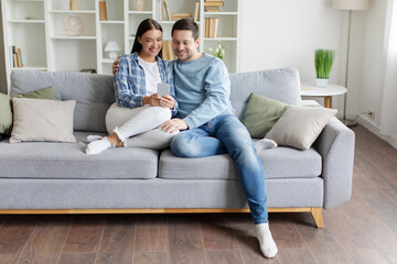 Young spouses enjoying leisure time using cellphone, embracing while sitting on couch in living room at home, full length, free space. Modern communication concept